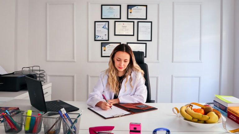Nutritionist sits at a modern office desk writing, with fruits and diplomas displayed, emphasizing health and professionalism.