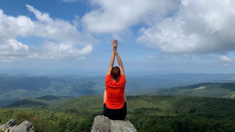 A woman in a red shirt relaxes on a mountain peak with arms raised, enjoying the scenic view.