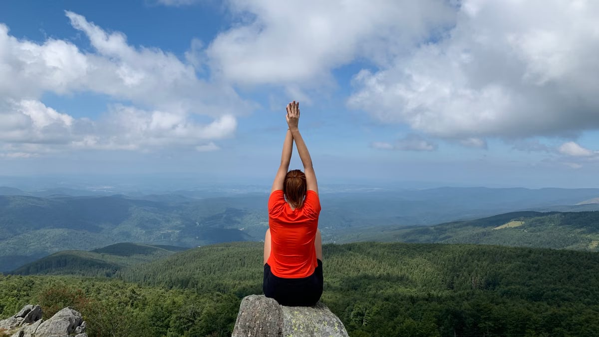 A woman in a red shirt relaxes on a mountain peak with arms raised, enjoying the scenic view.